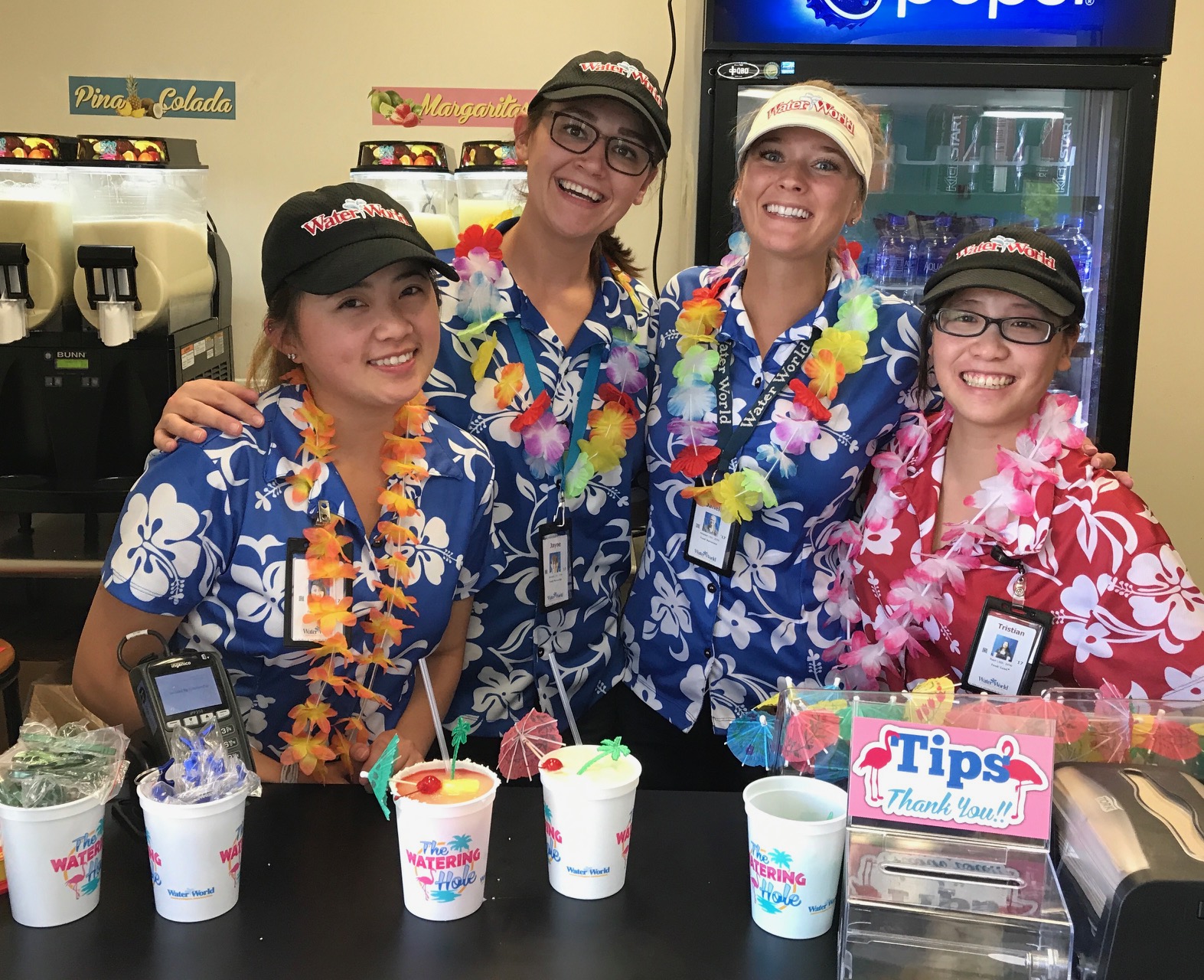 Four women in Hawaiian shirts and leis pose for a photo behind a food stand at Water World.